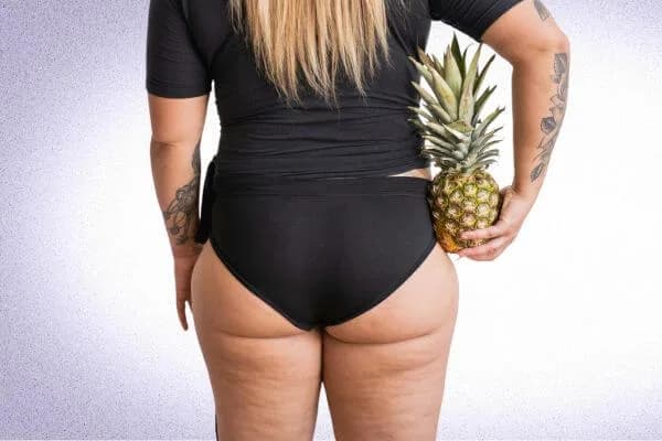 Path to empowerment: Woman wearing black underwear, embracing her body and showcasing cellulite on the back of her thighs and buttocks while holding a pineapple against a white background.